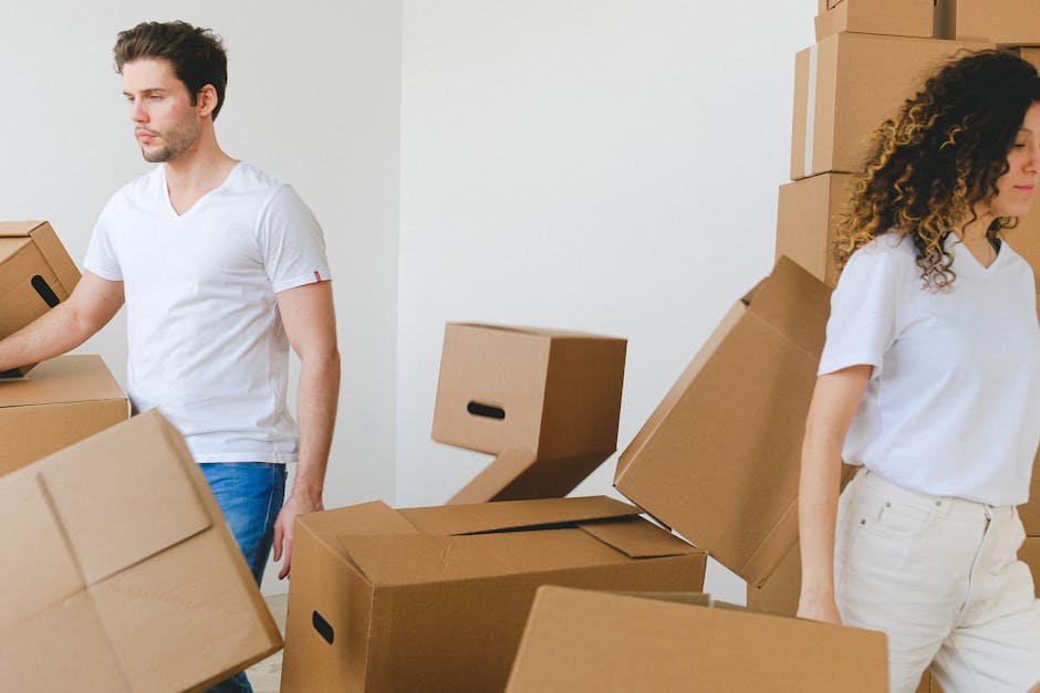 Two individuals involved in a home relocation, packing and moving process within an indoor space. The man on the left, dressed in a white t-shirt and blue jeans, is handling a large cardboard box, pos