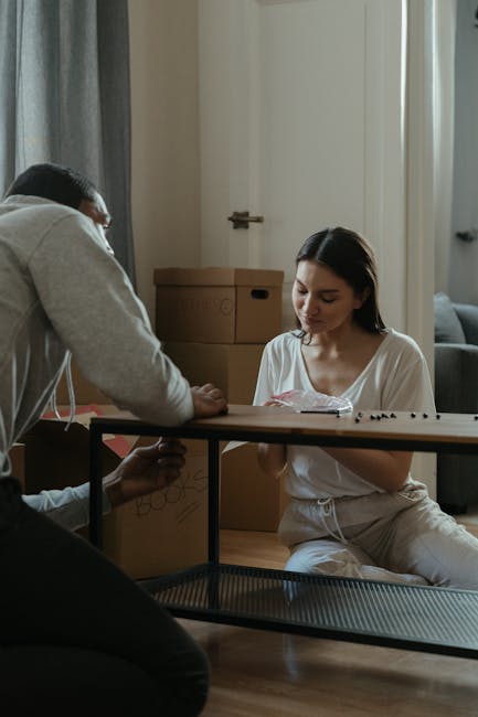 Two individuals involved in a home relocation, packing and moving process within an indoor space. The man on the left, dressed in a white t-shirt and blue jeans, is handling a large cardboard box, pos